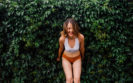 An example of boudoir photography where a woman in a white bra and red/orange underwear leans down, laughing, with her back to a beautiful green wall of foliage