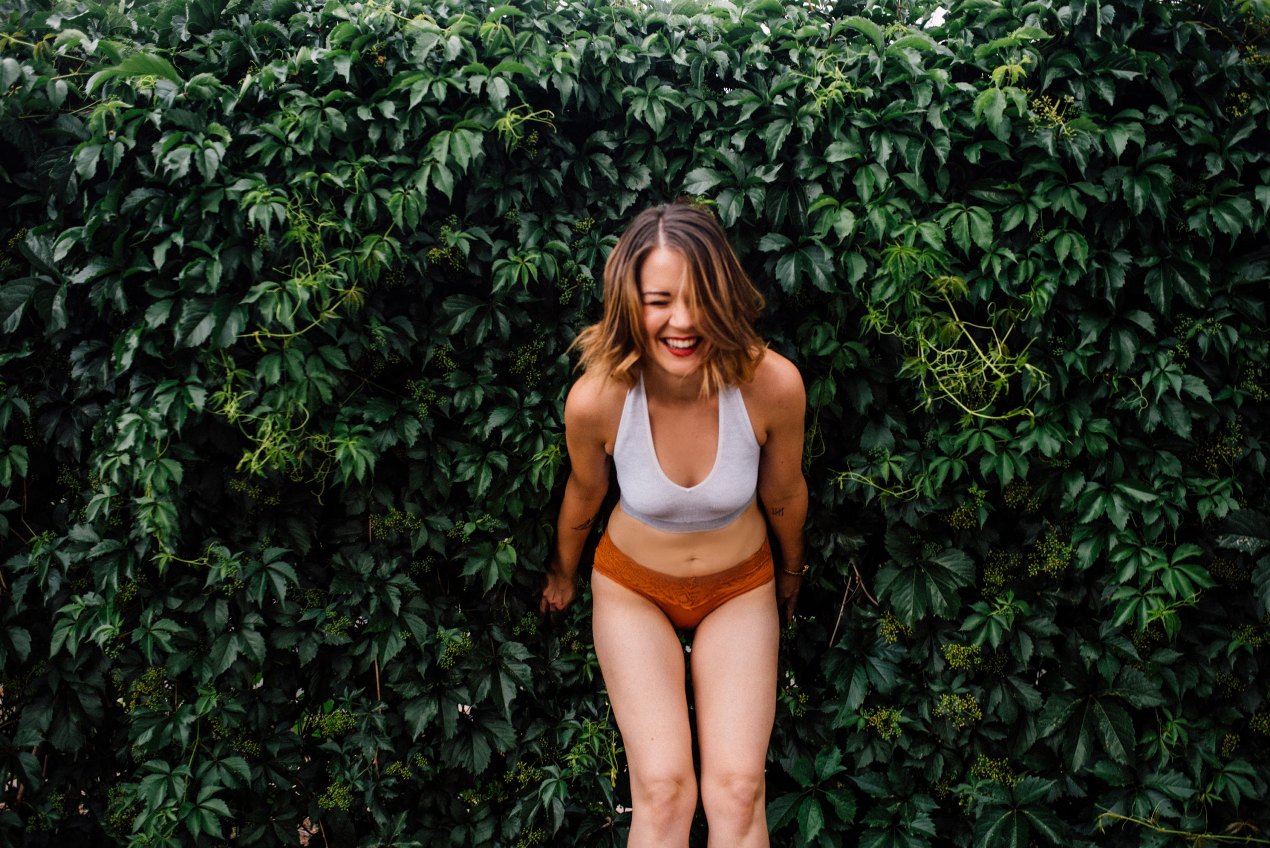 An example of boudoir photography where a woman in a white bra and red/orange underwear leans down, laughing, with her back to a beautiful green wall of foliage