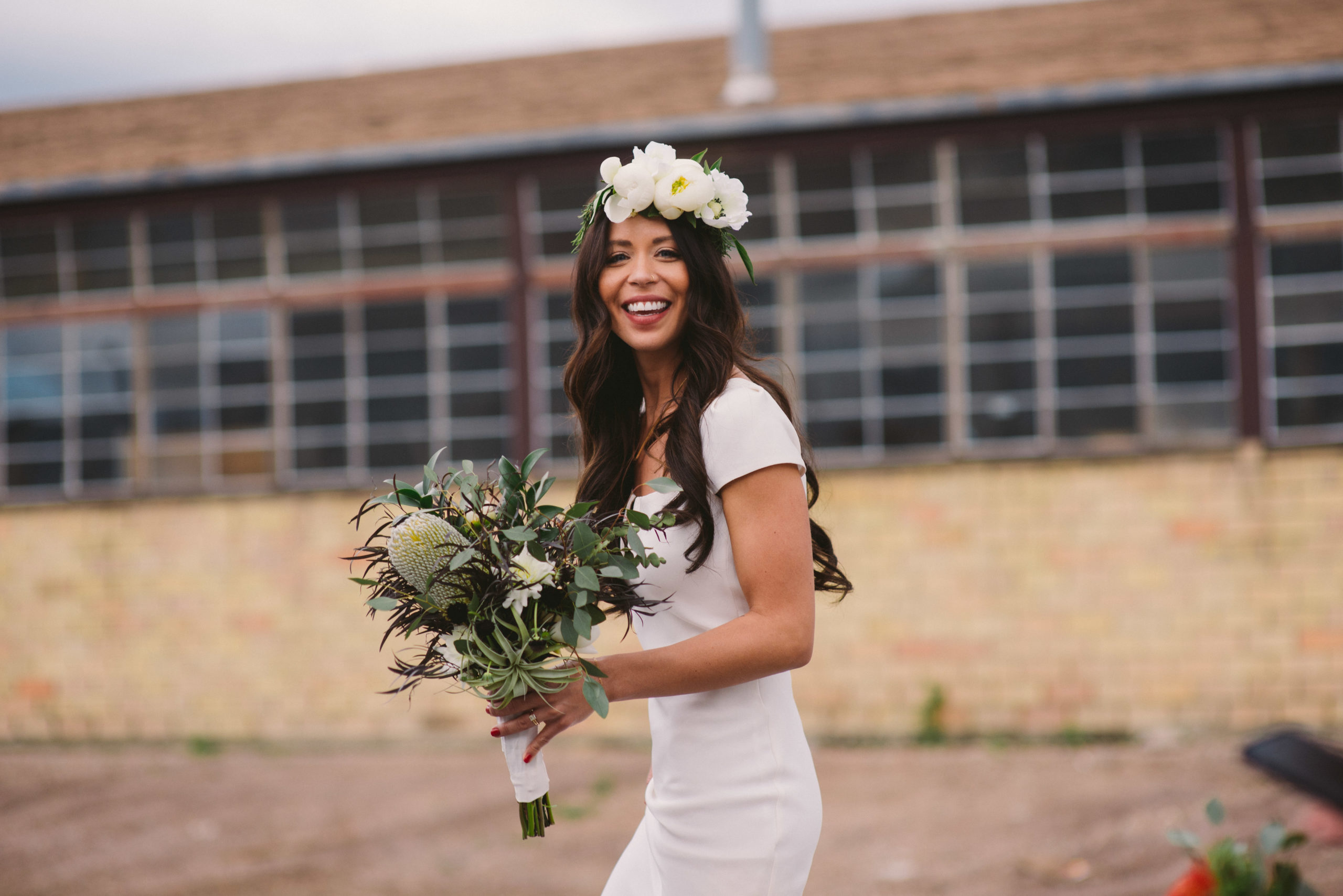wedding photography of bride looking towards camera with big smile wearing a flower crown and holding a big bouquets of green flowers casually walking by an old factory window