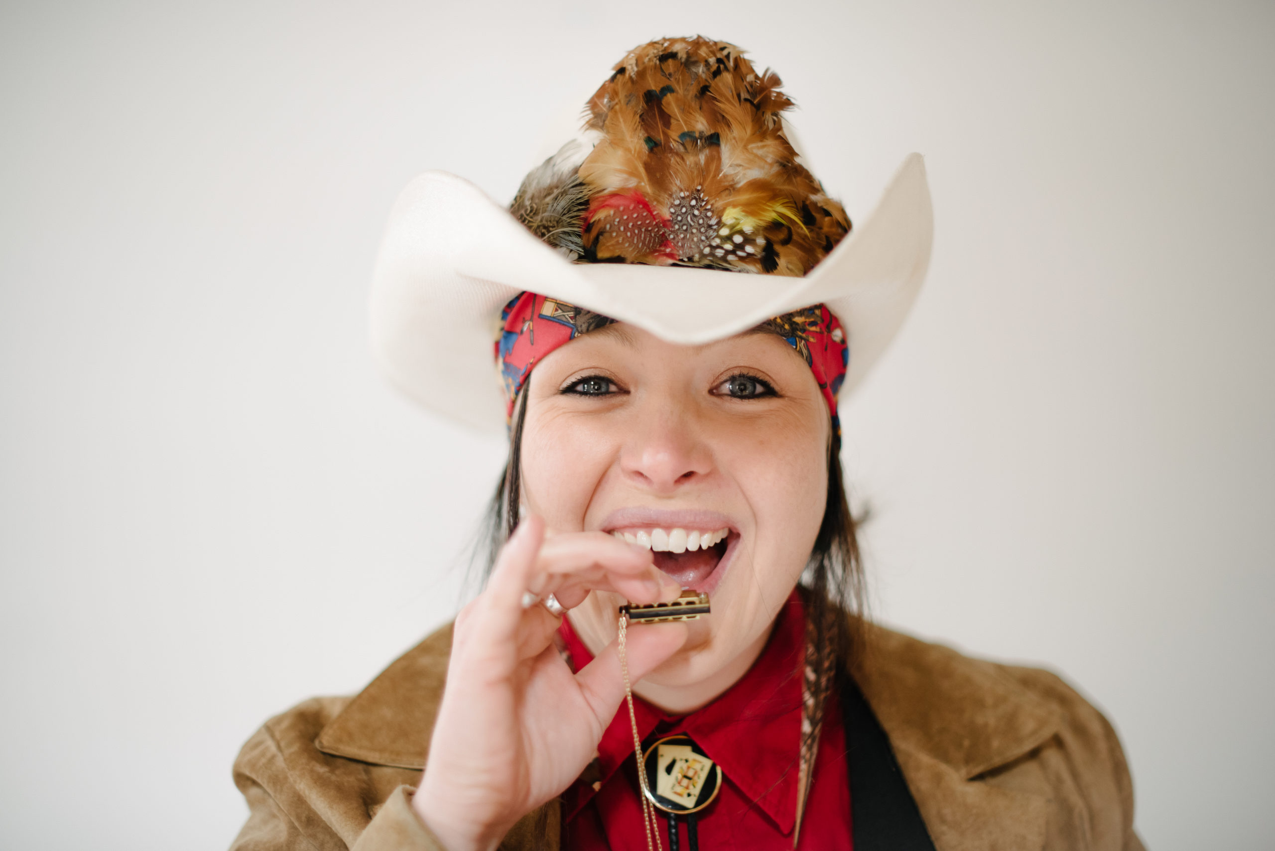 musician wearing large western hat with feathers and a bandana playing a tiny harmonica attached to a chain around her neck