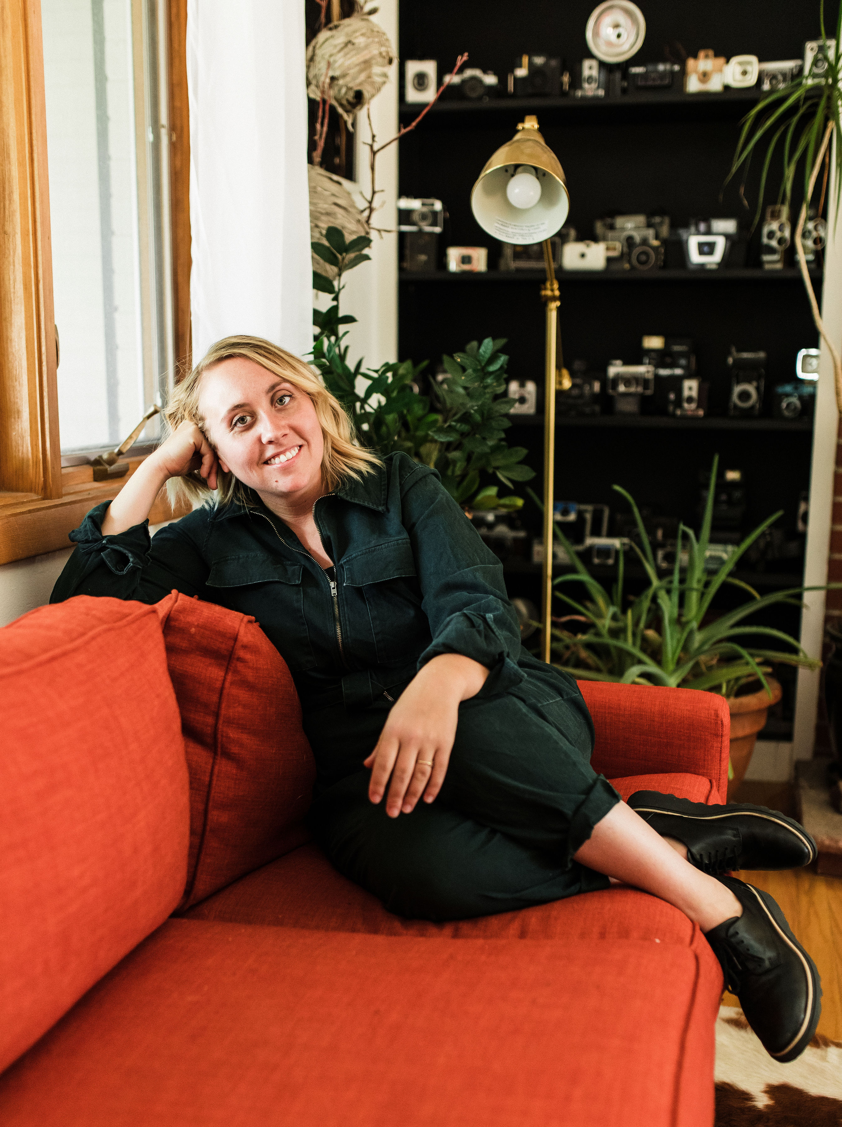 blonde girl leaning on the back of orange couch with her feet up wearing a dark green jumpsuit with a bookcase of old cameras in the background