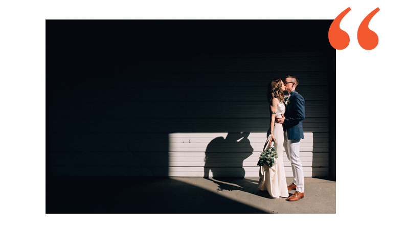 lesbian couple standing in a pocket of light in an alleyway. one is wearing a sleek off white wedding dress and the other is wearing a blue suit jacket with white pants and brown shoes.