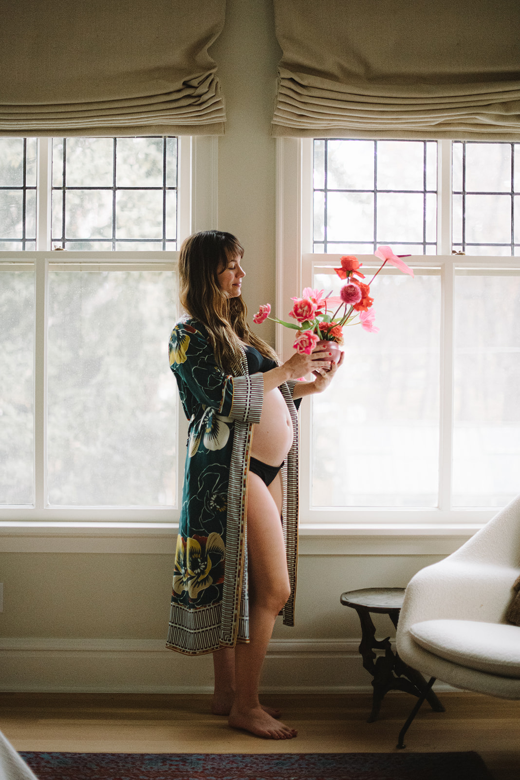 pregnant woman wearing only a robe standing in a window holding a bouquet of pink flowers