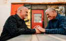 two older men with grey hair sitting across the table from each other holding hands smiling