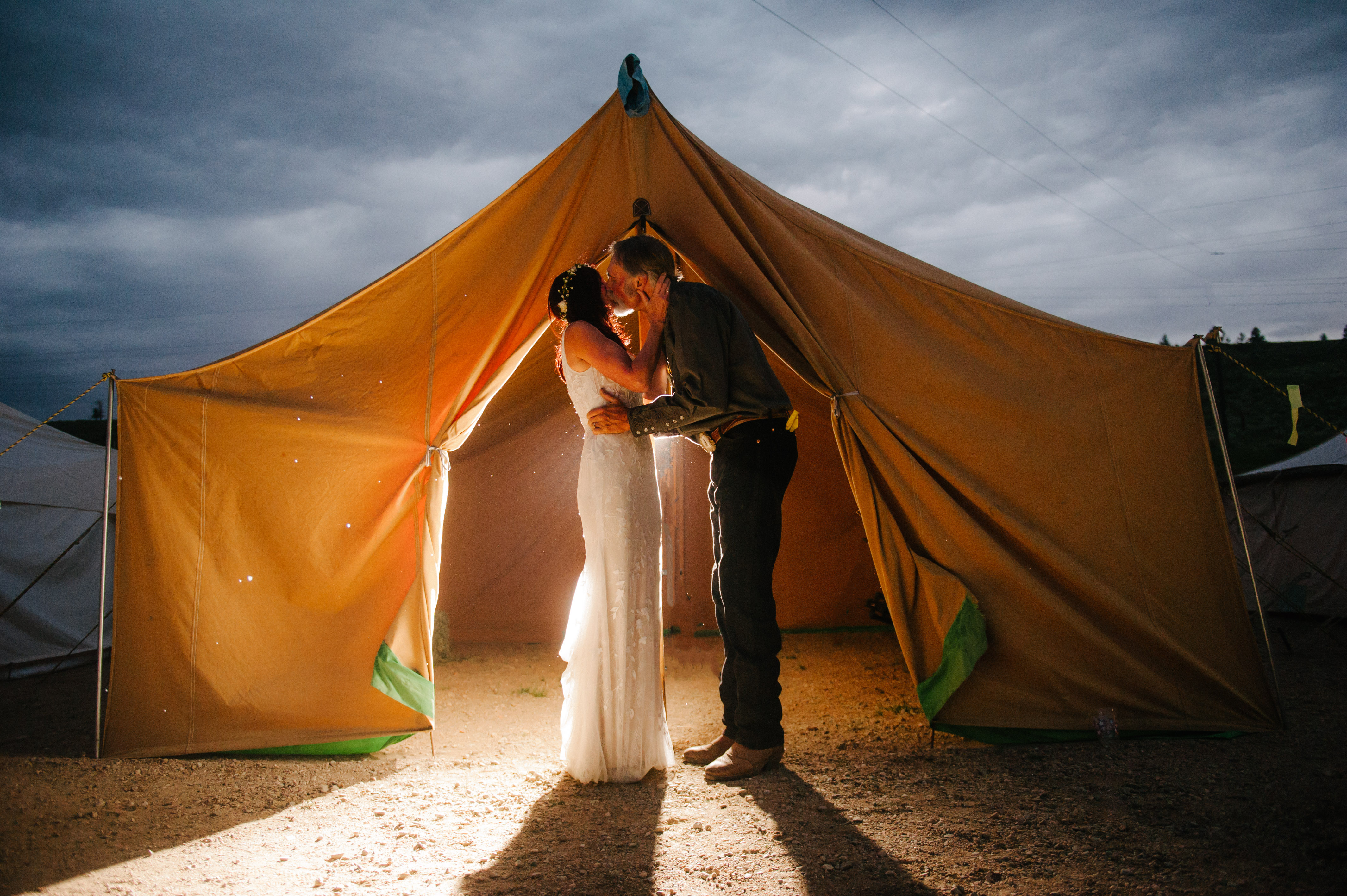 wedding photograph taken at dusk of a bride and groom kissing in front of a small tent that is illuminated by a flash coming from within